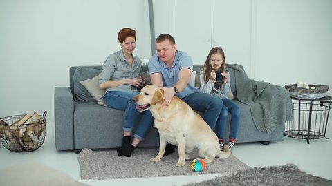Family of Three Playing with Dog at Home. Woman and her Daughter Holding Cups with Tea or Coffee in Hands while Sitting on the Sofa. Slow Motion. Pets and Animals Concept