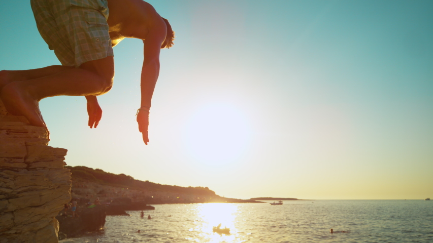 SLOW MOTION, CLOSE UP, LENS FLARE: Fit male tourist dives into the refreshing ocean water at sunset. Athletic man on summer vacation in Croatia dives off a rocky cliff on a perfect summer evening. Royalty-Free Stock Footage #1049462173