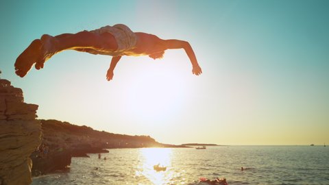 SLOW MOTION, CLOSE UP, LENS FLARE: Fit male tourist dives into the refreshing ocean water at sunset. Athletic man on summer vacation in Croatia dives off a rocky cliff on a perfect summer evening.