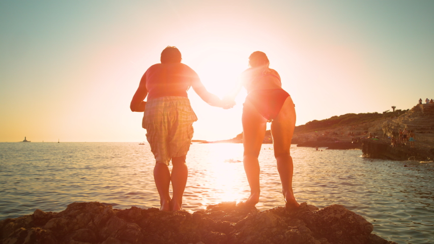 SLOW MOTION, LOW ANGLE, LENS FLARE: Cheerful couple on summer vacation jumps into the ocean at sunrise. Golden evening sun rays shine on young man holding girlfriend's hand as they jump in the water. | Shutterstock HD Video #1049462176