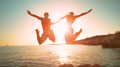 SLOW MOTION, LOW ANGLE, LENS FLARE: Cheerful couple on summer vacation jumps into the ocean at sunrise. Golden evening sun rays shine on young man holding girlfriend's hand as they jump in the water.