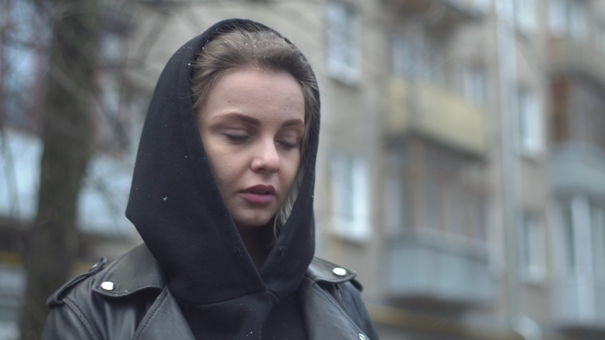 Young blonde in a black hood and leather jacket walks a dog on a leash in the snow on the street