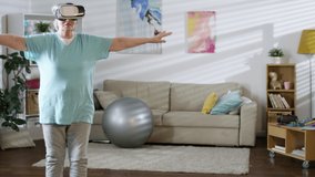 Senior woman in VR glasses doing standing front leg raise exercise while doing fitness in the living room at home