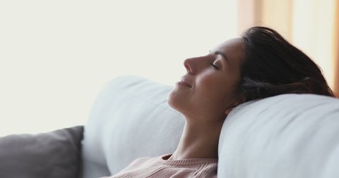 Healthy serene young woman leaning on cozy sofa taking deep breath of fresh clean air. Calm lady relaxing on comfortable couch, napping, enjoying no stress concept at home, close up side face view.