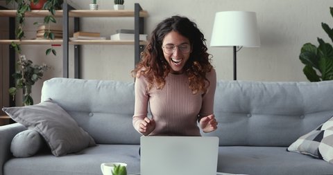 Excited young woman winner looks at laptop celebrates online success sits on sofa at home. Euphoric lady gets new distance job opportunity, reads good news in email, rejoices victory, feels motivated.