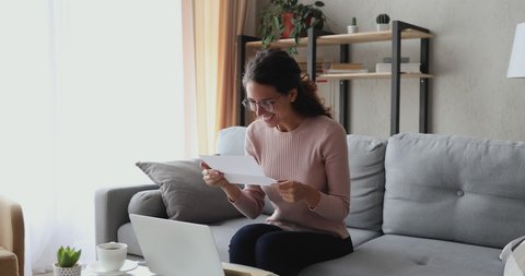 Happy excited woman reads good news in mail letter sits on sofa at home. Overjoyed lady winner holds bank paper receiving approved loan, tax refund or salary notice, celebrating success concept.