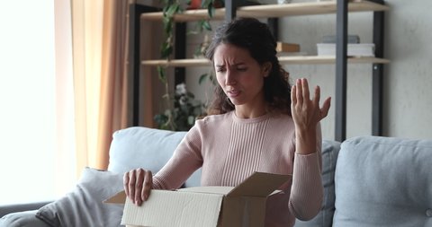 Frustrated upset young woman customer unpacking delivery at home. Angry dissatisfied shopper opening cardboard box feels bad surprise about damaged wrong bad parcel. Postal shipping problem concept