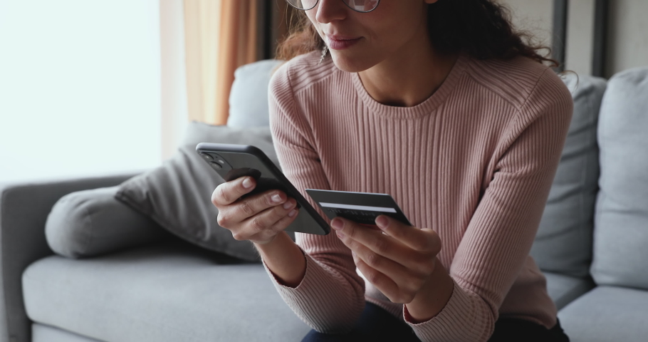 Smiling young woman customer holding credit card and smartphone sitting on couch at home. Happy female shopper using instant easy mobile payments making purchase in online store. E-banking app service | Shutterstock HD Video #1049471848