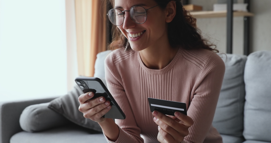 Smiling young woman customer holding credit card and smartphone sitting on couch at home. Happy female shopper using instant easy mobile payments making purchase in online store. E-banking app service