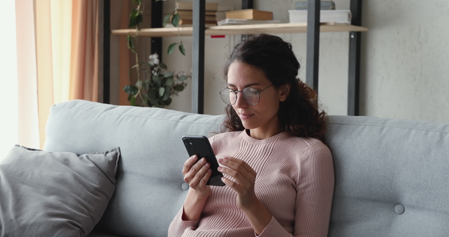 Relaxed young woman using smart phone surfing social media, checking news, playing mobile games or texting messages sitting on sofa. Millennial lady spending time at home with cell gadget technology. | Shutterstock HD Video #1049471884