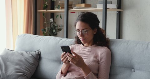 Relaxed young woman using smart phone surfing social media, checking news, playing mobile games or texting messages sitting on sofa. Millennial lady spending time at home with cell gadget technology.