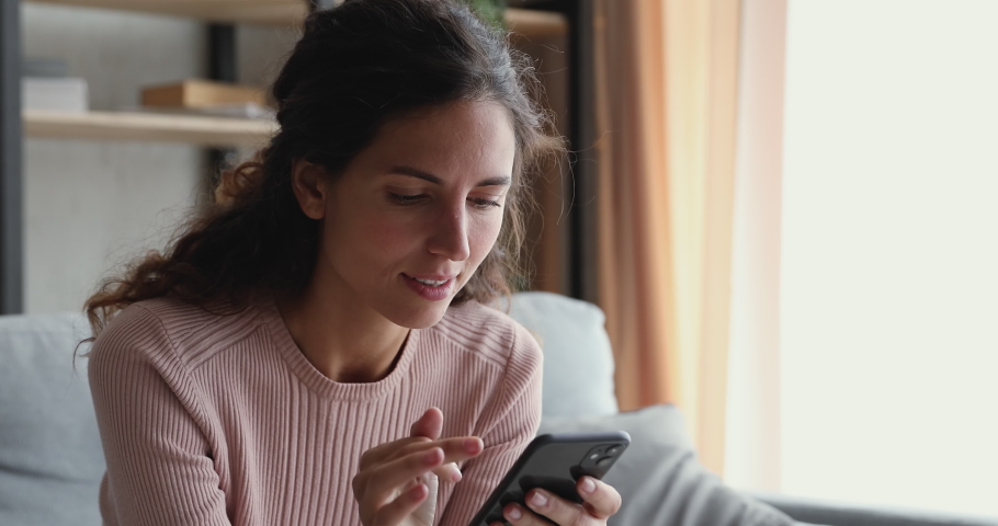Smiling millennial woman watching funny video stories on smart phone at home. Happy female customer doing mobile shopping, reading news, using apps, surfing internet looking at cell sitting on couch. Royalty-Free Stock Footage #1049471896
