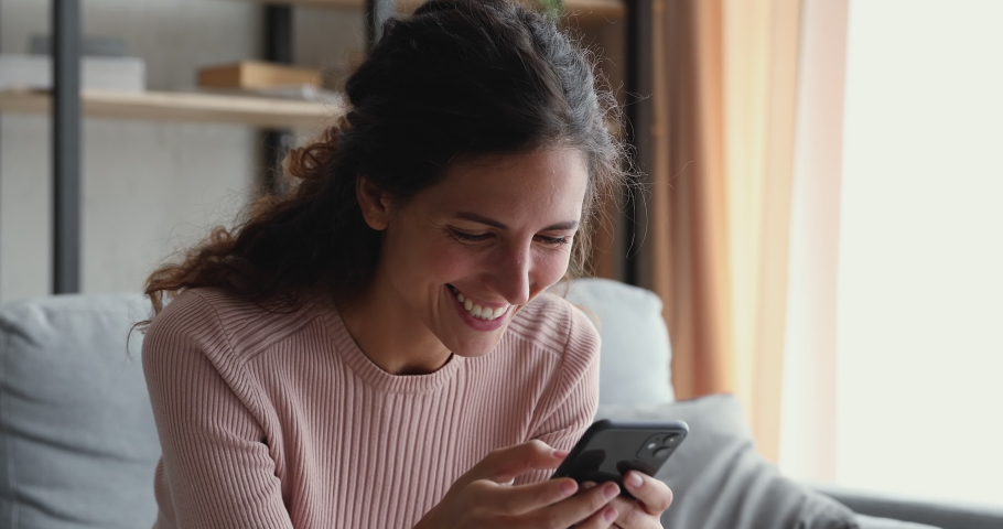 Smiling millennial woman watching funny video stories on smart phone at home. Happy female customer doing mobile shopping, reading news, using apps, surfing internet looking at cell sitting on couch. Royalty-Free Stock Footage #1049471896