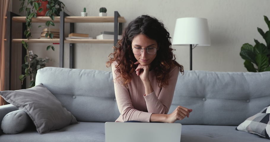 Millennial woman using laptop computer working online sitting on sofa at home office. Smart young lady doing online job thinking of internet research, browsing web, e learning with pc and papers.
