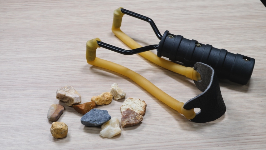 Slingshot small hand-powered projectile weapon. Classic form Y-shaped frame held in off hand with two rubber strips attached to uprights. Other ends of strips lead back to pocket that holds projectile