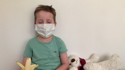 A small boy in a protective mask from viruses and diseases, eating a banana, against the background of a Teddy bear.