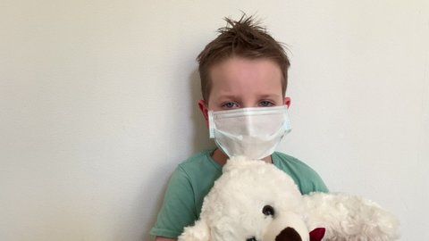 A child with a protective mask sits with a Teddy bear, at home in quarantine. looking at the camera, blinking.