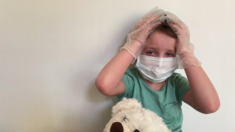 A crying child in a mask and latex gloves with a Teddy bear looks at the camera.