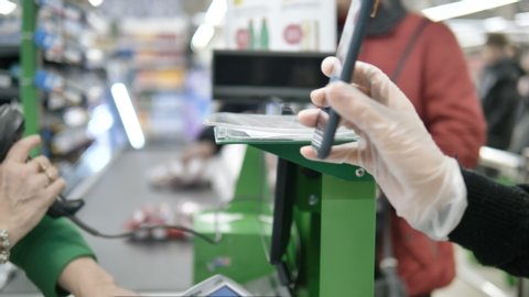 A woman at the checkout in a supermarket gives a customer a card with a laser from her smartphone. Paying in the supermarket in protective gloves so as not to become infected with a coronavirus