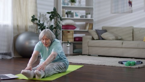 Tracking shot of senior woman watching video workout on laptop and doing stretching exercise on mat in the living room while exercising at home