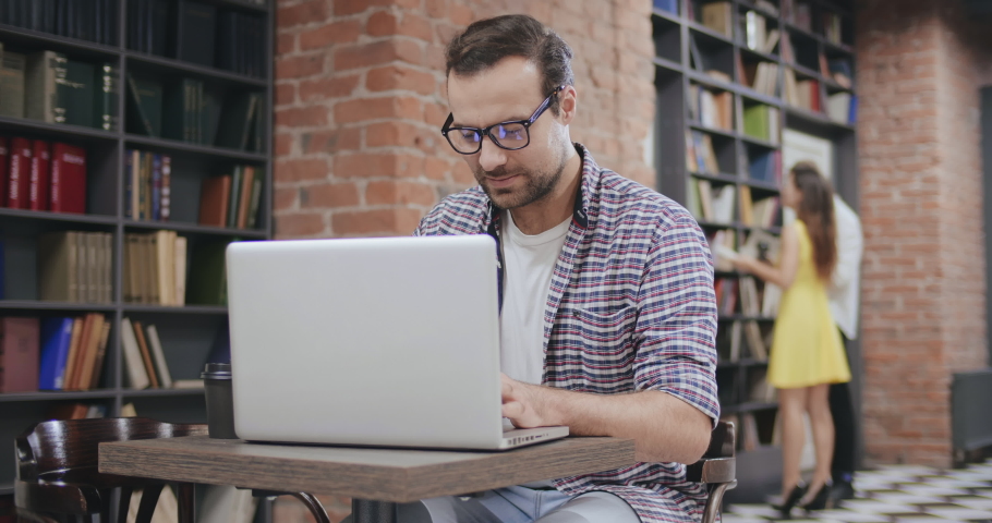 Portrait of young man in glasses using laptop while studying in college library with other people near bookshelves on background. Man working on laptop in book cafe Royalty-Free Stock Footage #1049479489