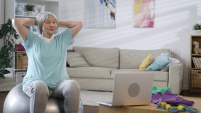 Senior woman holding hands behind head and moving on stability ball while following workout lesson on laptop in the living room