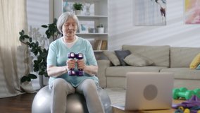 Cheerful senior woman sitting on stability ball, watching video workout on laptop and exercising with dumbbells in the living room at home