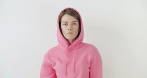 Portrait of young playful girl dressed in pink hoodie standing over white background blowing bubblegum candy bubbles and chewing gum. Woman looking at camera. 4k raw video footage slow motion