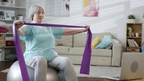 Senior woman sitting on stability ball, watching workout lesson on laptop and doing chest stretch exercise with resistance band while training at home