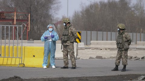 Almaty, Kazakhstan - March 20, 2020: Two soldiers in medical masks with a medic in a protective suit are standing at the checkpoint of a quarantined city