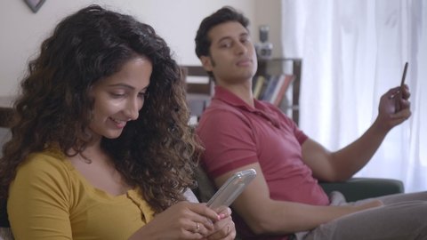 A young woman or wife is typing a text message on her mobile or smartphone or cellphone. Insecure couple sitting on a couch while a man is trying to have a peek into the girl's phone. 