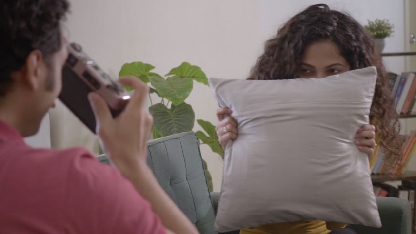 A shy woman is on a couch hiding behind a small pillow cushion while her husband is clicking a picture with a Vintage DSLR camera. A happy smiling couple enjoying the time together in the house | Shutterstock HD Video #1049496337