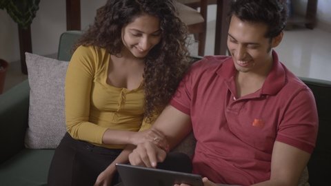 A young attractive good looking couple watching an online comedy movie on a tablet while sitting on a couch in the house. Husband and wife laughing smiling together indoors. relationship concept