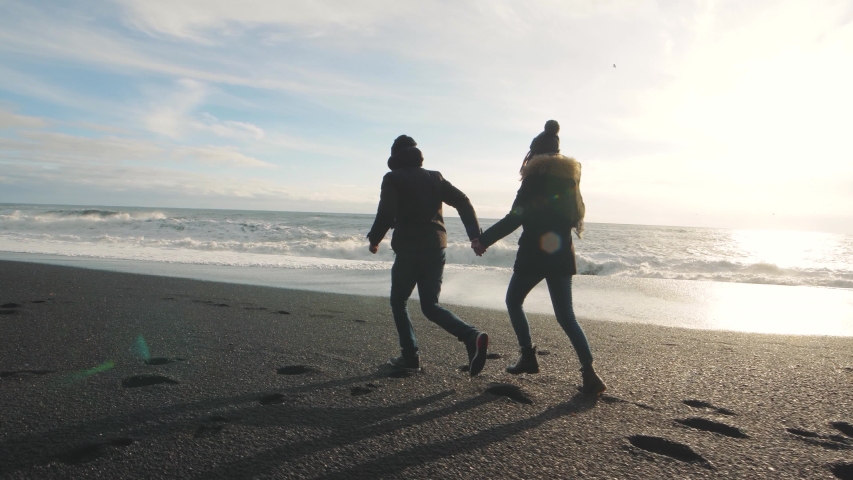 Couple in love running on volcanic black sand beach in Iceland and having fun at sunset, slow motion. Reynisfjara Beach, Reynisdrangar troll toes mountains, Vik. Iceland in winter Royalty-Free Stock Footage #1049501965