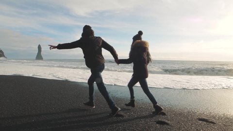 Couple in love running on volcanic black sand beach in Iceland and having fun at sunset, slow motion. Reynisfjara Beach, Reynisdrangar troll toes mountains, Vik. Iceland in winter