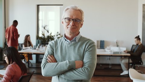 Waist up shot of senior caucasian male manager in glasses and casual outfit looking at camera and smiling while posing with arms crossed in open space office during workday