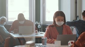Mixed raced woman in protective face mask sitting at desk and typing on laptop while working in open space office with colleagues during coronavirus outbreak - Powered by Shutterstock - Get 15% off with code: PIKWIZARD15