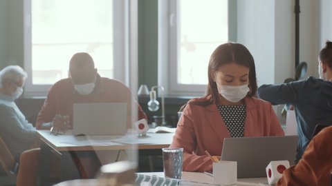 Mixed raced woman in protective face mask sitting at desk and typing on laptop while working in open space office with colleagues during coronavirus outbreak
