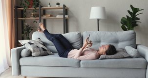 Relaxed young woman lying on sofa in living room using smart phone. Lazy happy millennial lady customer surfing social media, shopping in app, playing mobile game, texting sms looking at cell at home.