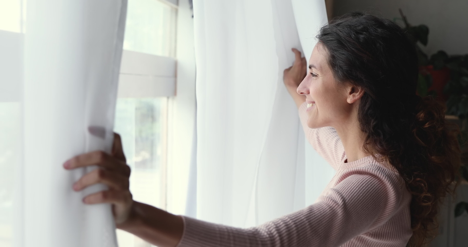 Happy optimistic young woman opening curtain lace looking through window. Smiling confident lady enjoying watching beautiful cityscape view, feeling hope and dreaming of good future at home. | Shutterstock HD Video #1049522455