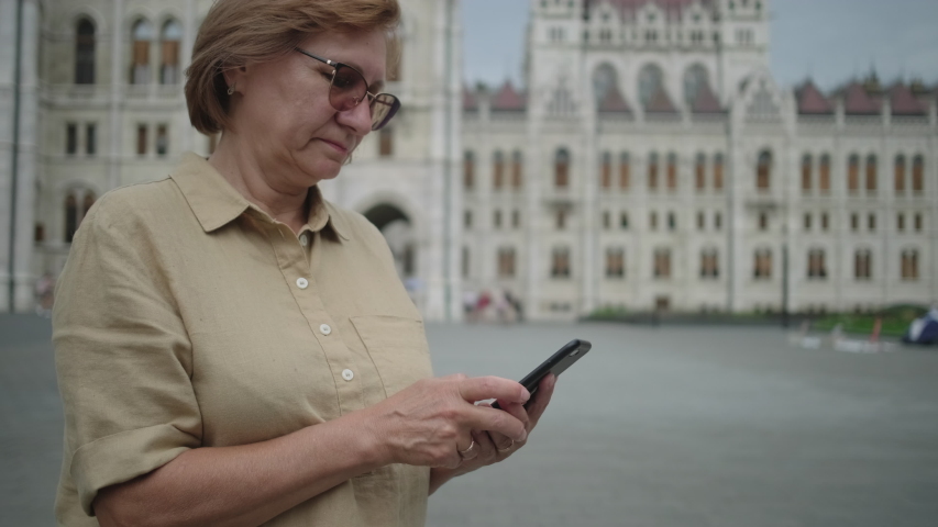 Senior woman in glasses chatting on smartphone at Kossuth Square with Hungarian Parliament Building in Budapest. Notable city landmark
