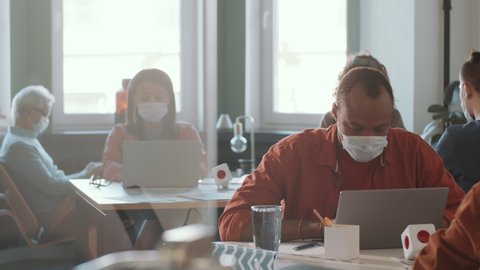 Young African American man in protective face mask sitting at table and using laptop while working in open space office with diverse colleagues during coronavirus outbreak Stock Video