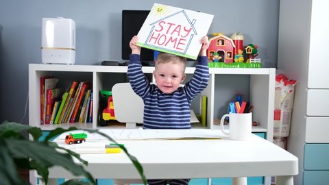 Coronavirus covid-19 slogan stay home. Caucasian boy in home self-isolation holds inscription "stay home" hides his head and looks out from behind paper. Coronavirus don't leave your house on street