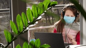 teenager girl in mask and headphones, listening to music on laptop, chatting online, on open balcony. spring sunny day. quarantine. stay, study at home. coronavirus epidemic.