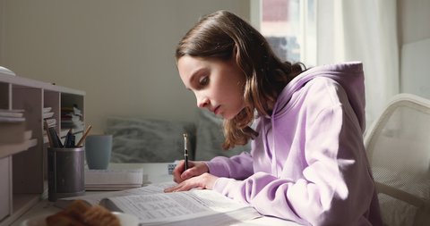 Distance learning concept. Adolescent teen girl studying, reading textbook, making notes in copybook sitting at home desk. Smart teenage school student learning alone doing homework assignment.