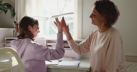 Happy family adult parent mother helping motivating teenage school girl doing homework giving high five. Positive mom and teen daughter studying together having fun sitting at home desk in sunny room.