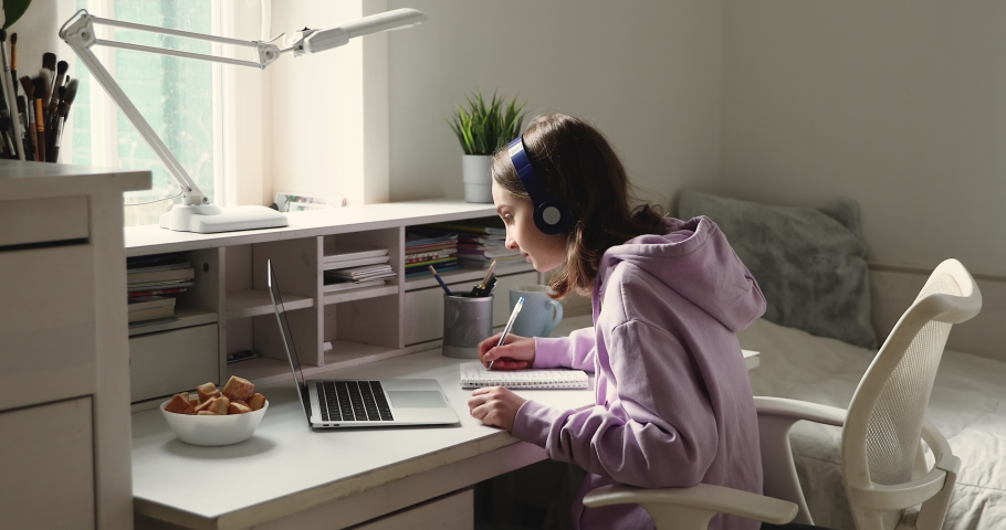 Distance learning concept. Adolescent schoolgirl studying online using laptop making notes in copybook. Teen girl school student wearing headphones watching internet video course sitting at home desk. | Shutterstock HD Video #1049543275