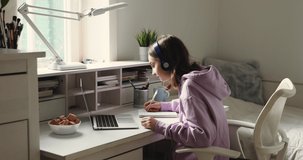 Distance learning concept. Adolescent schoolgirl studying online using laptop making notes in copybook. Teen girl school student wearing headphones watching internet video course sitting at home desk.