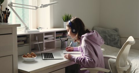 Distance learning concept. Adolescent schoolgirl studying online using laptop making notes in copybook. Teen girl school student wearing headphones watching internet video course sitting at home desk.