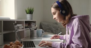 Teenage girl wearing headphones studying online from home with distance teacher. Teen school student distance learning in internet making notes, doing homework, watching video tutorial course concept.